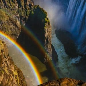 Un doppio arcobaleno si inarca su una gola profonda e rocciosa con una grande cascata sullo sfondo sotto un cielo sereno.