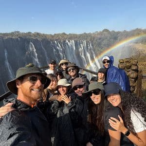 Un gruppo WeRoad si fa un selfie con attrezzatura antipioggia di fronte a una grande cascata con un arcobaleno nel cielo.