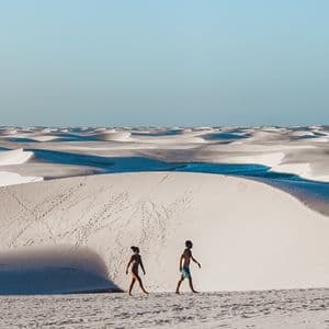 Deux personnes en maillot de bain traversent de vastes dunes de sable blanc avec des lagons bleus sous un ciel clair.