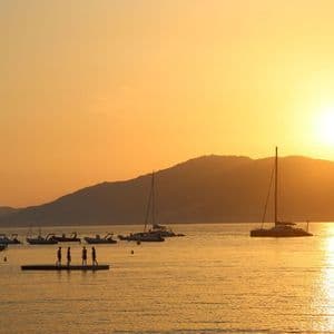 Silhouettes d'un groupe WeRoad en voyage sur un ponton flottant dans une baie, avec des bateaux, au coucher du soleil derrière les collines.