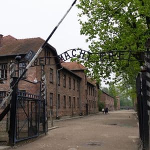 El letrero 'Arbeit Macht Frei' sobre la puerta de entrada principal de un antiguo campo de concentración, con barracones de ladrillo bordeando un camino de tierra.