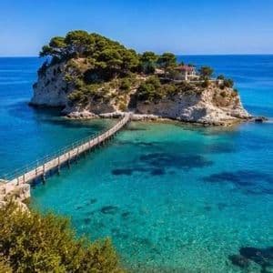 Un ponte di legno si estende su acque cristalline color turchese fino a una piccola isola rocciosa, coperta di alberi, sotto un cielo azzurro brillante.