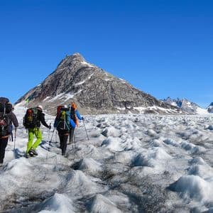 Un viaggio di gruppo WeRoad con tre escursionisti che, muniti di zaini, fanno trekking su un vasto ghiacciaio ondulato, diretti verso una montagna rocciosa sotto un cielo azzurro e limpido.