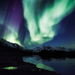 L'aurora boreale verde e viola brilla nel cielo notturno, riflettendosi su un lago calmo e montagne innevate.
