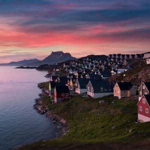 Un borgo costiero con case colorate è illuminato lungo una costa rocciosa al tramonto, con un cielo vibrante di rosa e viola.
