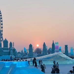 Un'ampia vista di un festival invernale con persone tra grandi sculture di ghiaccio e una ruota panoramica contro un cielo al tramonto.