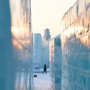 Una persona cammina su un sentiero innevato tra imponenti pareti di ghiaccio, con una pagoda di ghiaccio visibile in lontananza al tramonto.