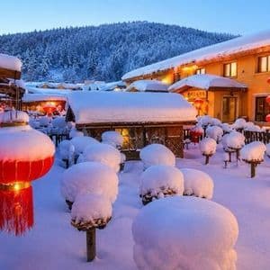 Un villaggio innevato al crepuscolo, con lanterne rosse accese, edifici illuminati da una luce calda e lo sfondo di una foresta imbiancata.