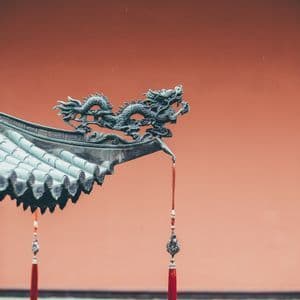 Un primo piano di una scultura di drago in metallo sulla grondaia di un tetto in tegole, con una nappa rossa pendente contro un muro rosso.