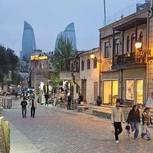 Menschen spazieren in der Dämmerung durch eine gepflasterte Straße mit Geschäften, während im Hintergrund moderne Wolkenkratzer zu sehen sind.