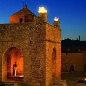 Ein Steintempel mit Feuern auf dem Dach und einer Feuerstelle im Innenhof bei Dämmerung, mit einer Person, die in einem Torbogen steht.