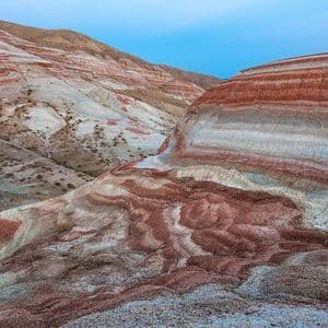 Schichten aus rotem, weißem und grauem Sediment bilden sanfte, gestreifte Hügel unter einem hellblauen Himmel.