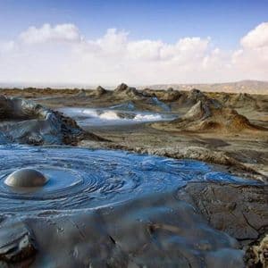 Ein blubbernder Schlammvulkan erzeugt konzentrische Wellen in einer ariden Landschaft mit zahlreichen kleineren Schlammhügeln unter bewölktem Himmel.