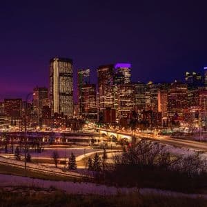 Une ligne d'horizon urbaine de gratte-ciel illuminés, sous un ciel de crépuscule pourpre, avec un sol enneigé et un pont éclairé au premier plan.