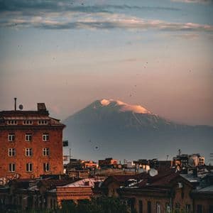 Une grande montagne enneigée domine les toits de la ville et la cime des arbres verts sous un ciel de coucher de soleil nuageux.