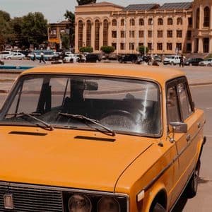 Une voiture ancienne jaune garée sur une place de ville devant un grand bâtiment classique avec des gens en arrière-plan.