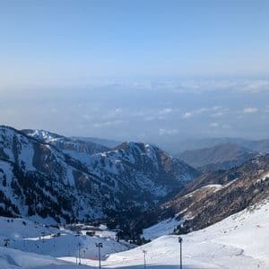 Vue plongeante d'une vallée de montagne enneigée avec des skieurs sur une piste sous un ciel bleu dégagé.