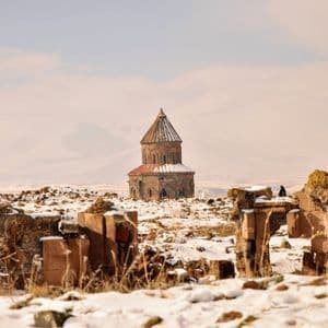 Une ancienne église en pierre au toit conique se dresse dans un vaste paysage enneigé, vue de derrière des ruines délabrées.