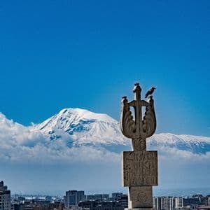 Un oiseau noir est perché sur un monument en pierre sculptée surplombant une ville, avec une montagne enneigée s'élevant au-dessus des nuages.