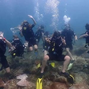 A WeRoad group trip of scuba divers posing together underwater above a rocky, coral-covered seabed, making hand gestures.