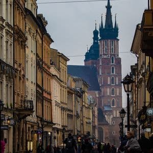 Des gens marchent dans une rue pavée et humide, bordée de bâtiments historiques, menant à une grande église en briques avec une tour proéminente.