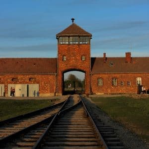 Des rails de chemin de fer mènent vers l'entrée voûtée d'un bâtiment en briques rouges, doté d'une tour de guet, sous un ciel bleu au crépuscule.