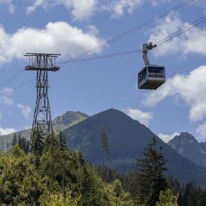 Un téléphérique circule sur ses câbles au-dessus d'une forêt verdoyante, avec un grand sommet de montagne visible sur un ciel partiellement nuageux.
