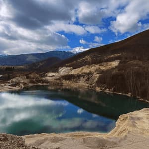 Un lago calmo in una cava riflette un cielo azzurro nuvoloso, con colline brulle e lontane montagne innevate sullo sfondo.