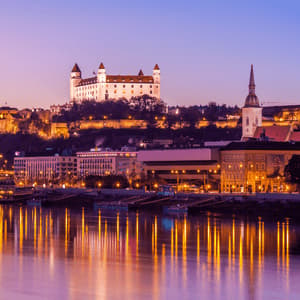 Un castello bianco illuminato su una collina domina lo skyline di una città vicino a un fiume al tramonto, con le luci che si riflettono nell'acqua.