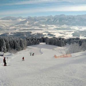 Sciatori su un'ampia pista innevata vicino a uno chalet, con vista su una pineta e una vasta valle di montagna.