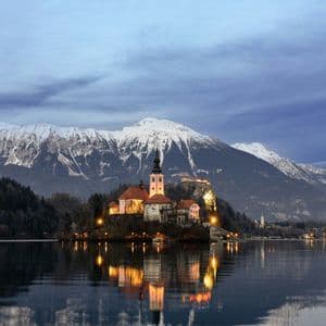 Una chiesa illuminata su una piccola isola si riflette nelle acque calme di un lago al tramonto, con montagne innevate sullo sfondo.
