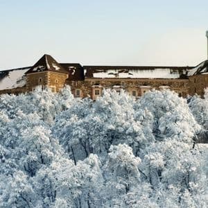 Un castello in pietra con una torre dell'orologio si erge sopra una fitta foresta di alberi completamente coperti di neve bianca.