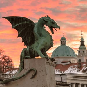 Una grande statua di drago alato in bronzo si erge su un piedistallo di pietra, stagliandosi contro un cielo colorato al tramonto su una città con cupole.
