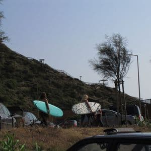 Un viaggio di gruppo WeRoad trasporta tavole da surf attraverso un parcheggio con una grande collina verde sullo sfondo.