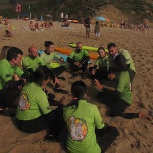 Un gruppo WeRoad, che indossa top verdi e mute abbinate, è seduto in cerchio su una spiaggia sabbiosa con le tavole da surf.