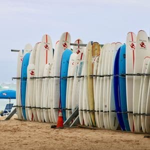 Una grande rastrelliera di tavole da surf bianche e blu si trova su una spiaggia sabbiosa, con le onde dell'oceano visibili sullo sfondo.