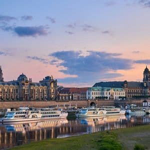 Un panorama storico cittadino con cupole e guglie ornate, che si riflette in un fiume al tramonto, con barche turistiche ormeggiate lungo la riva.