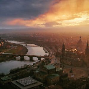 Veduta aerea di una città storica con un ampio fiume e un ponte ad arco, illuminata dalla calda luce di un tramonto dorato.