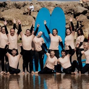 Un gruppo WeRoad in mute da sub, in posa per una foto su una spiaggia, sorridenti e che salutano con le loro tavole da surf blu.