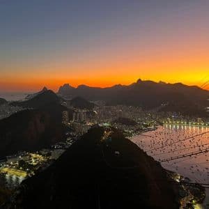 Vista dall'alto di una città costiera illuminata al tramonto, con montagne in silhouette e una baia piena di barche.