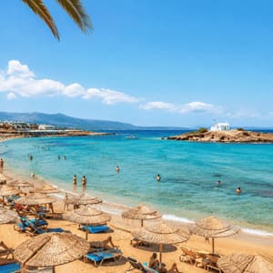 Una veduta dall'alto di una spiaggia sabbiosa affollata con ombrelloni di paglia, persone che nuotano nell'acqua turchese e una piccola isola con una cappella.