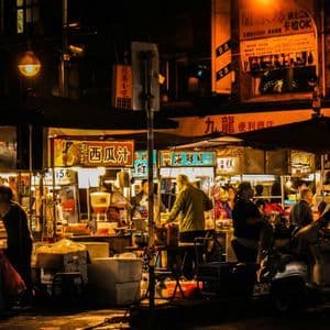 Un vivace mercato di strada notturno con persone riunite attorno a bancarelle di cibo illuminate e insegne in caratteri asiatici.