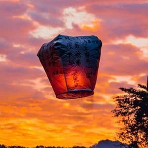 Una lanterna di carta illuminata con calligrafia fluttua in un cielo nuvoloso, arancione e viola al tramonto, sopra colline in silhouette.