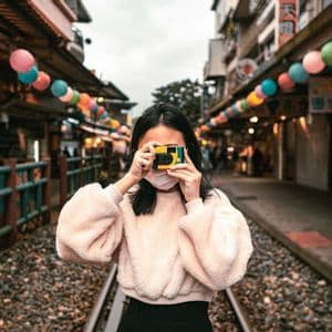 Una donna con la mascherina scatta una foto con una macchina fotografica, stando sui binari del treno in una strada di mercato con lanterne colorate.
