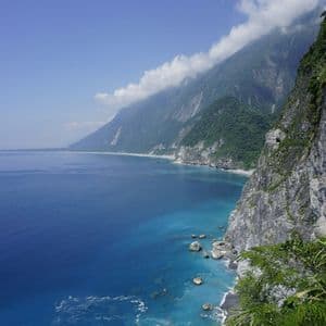 Una costa frastagliata dove ripide montagne verdi incontrano il mare vibrante blu e turchese sotto un cielo parzialmente nuvoloso.