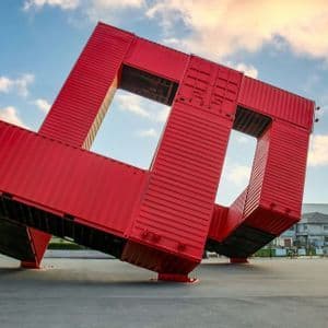 Una grande scultura rossa, fatta di container, è in equilibrio obliquo in una piazza pavimentata sotto un cielo blu con nuvole.
