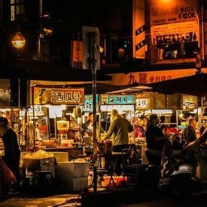 Un vivace mercato notturno asiatico, con persone radunate attorno a bancarelle di cibo illuminate sotto insegne luminose.