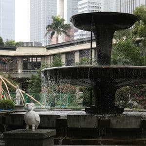 Una fontana scura a più livelli e una piccola statua di anatra bianca in un parco cittadino con moderni grattacieli sullo sfondo.