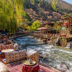 Un pintoresco café al aire libre con cojines y alfombras de colores se encuentra junto a un río caudaloso, con un pueblo en la ladera de fondo.