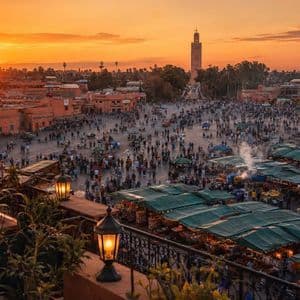 Vista elevada de una plaza concurrida con puestos de mercado y un minarete lejano bajo un atardecer naranja.
